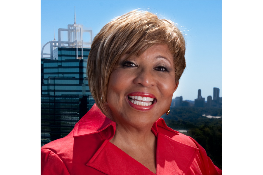 This is a Brian Charles Steel headshot of Mimi Williams.  The light on her face is soft.  She is wearing red lipstick and a red shirt with a collar.  Williams is positioned in the center of the frame, and composes about three fourths of the image.  She is smiling revealing her pearly white teeth.  The king and queen building are in the background in the left corner. 